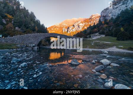 Malerischer Blick auf die Steinbrücke über den ruhigen Fluss mit steinigen Ufern, umgeben von Bergen und Bäumen bei Sonnenuntergang in den Pyrenäen Stockfoto