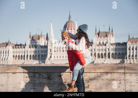 Seitenansicht eines romantischen glücklichen Mannes, der seiner Freundin in gemütlicher warmer Kleidung einen Huckepack gibt, während er sich zusammenhält und Zeit in Budapest, Ungarn, verbringt Stockfoto