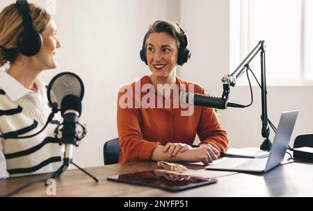 Zwei Frauen, die sich unterhalten, während sie eine Audioübertragung in einem Heimstudio moderieren. Zwei weibliche Content-Ersteller nehmen einen Internet-Podcast auf Stockfoto