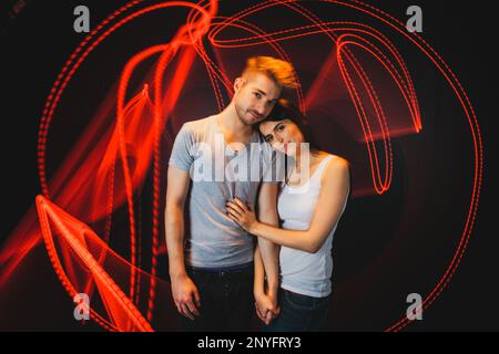 Netter Mann und Frau umarmen sich gegenseitig und betrachten Kamera im Stehen in der Nähe von hellen roten Lichtspuren Schwarzer Hintergrund Stockfoto