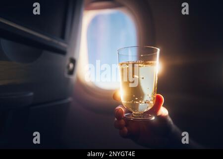 Ein Mann, der während des Fluges etwas trinkt. Passagier hält ein Glas Sekt gegen das Flugzeugfenster. Stockfoto