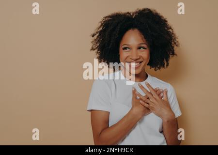 Porträt einer glücklichen afrikanerin in einem weißen T-Shirt, das auf beigefarbenem Hintergrund posiert, mit einem Lächeln, das wegschaut, Hände auf der Brust hält, fühlt sich Dankbarkeit und Geste an Stockfoto