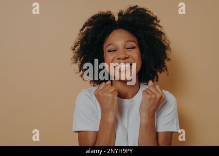 Überglücklich glücklich glücklich, dunkel gehäutete junge Frau erhebt geballte Fäuste, zeigt weiße Zähne, trägt legeres weißes T-Shirt, Models isoliert auf pastellbeigefarbenem Rücken Stockfoto