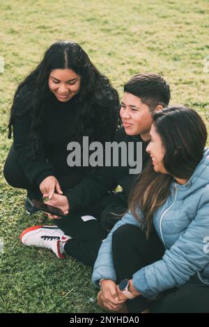 Drei Personen hispanisch-lateinamerikanischer Herkunft, die auf dem üppigen grünen Gras eines Parks sitzen, mit einem intelligenten Gerät. Entspannte und lächelnde Ausdrücke auf dem Stockfoto