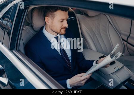 Der nachdenkliche junge, bärtige, gut aussehende Executive Manager im formellen dunkelblauen Anzug liest Zeitung auf dem Rücksitz der Limousine, fährt zur Arbeit in seinem Büro Stockfoto