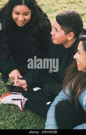 Drei Personen hispanisch-lateinamerikanischer Herkunft, die auf dem üppigen grünen Gras eines Parks sitzen, mit einem intelligenten Gerät. Entspannte und lächelnde hispanische Familie Stockfoto