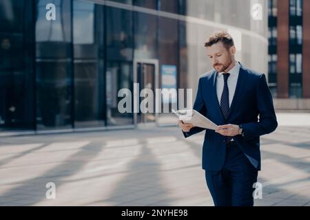 Fokussierter, bärtiger junger Geschäftsmann in formellen Outfits, der alleine mit einer Zeitung in den Händen spaziert und die neuesten Nachrichten auf dem Weg zur Arbeit mit Büroklamotten liest Stockfoto