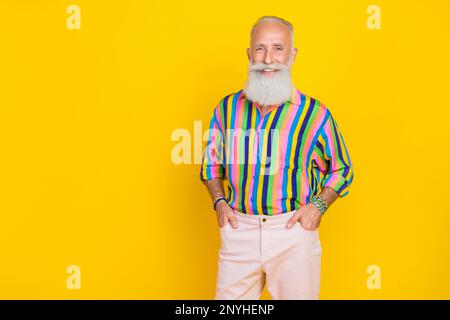 Porträt eines netten, gut aussehenden alten Mannes mit langem Bart Tragen Sie farbenfrohe Hemdarme in Taschen isoliert auf gelbem Hintergrund Stockfoto