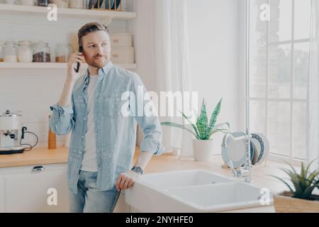 Junger Mann in lässigen Jeans Hemd, der mit seinem Partner am Handy spricht und wegschaut, während er an einem Holztisch in einer modernen Kücheneinrichtung steht Stockfoto