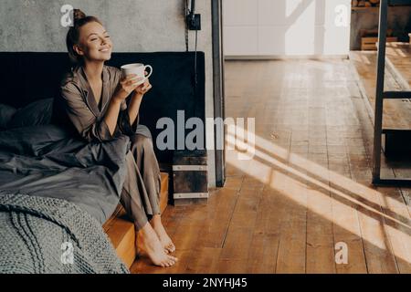 Ein neuer, schöner Tag. Süße hübsche Frau mit einer Tasse Kaffee am Morgen, die sich kühlt, während sie auf dem Bett sitzt und sich im braunen Satin-Pyjama im modernen Nebeneinander erholt Stockfoto