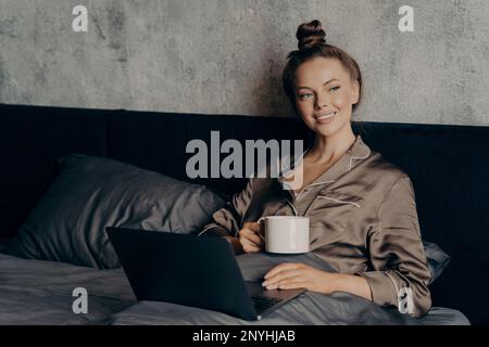 Faule, entspannte Morgenzeit. Lächelnde junge Frau, die im Bett im satinierten Pyjama lag, mit einer Tasse Kaffee in der Hand, während sie die letzten Nachrichten auf Inte überprüfte Stockfoto