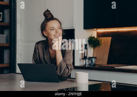 Junge, fröhliche Freiberuflerin in gemütlichen Pyjamas, die von zu Hause aus arbeitet und am Morgen ihre E-Mails auf ihrem Laptop abliest, während sie eine Tasse Kaffee im Stil trinkt Stockfoto