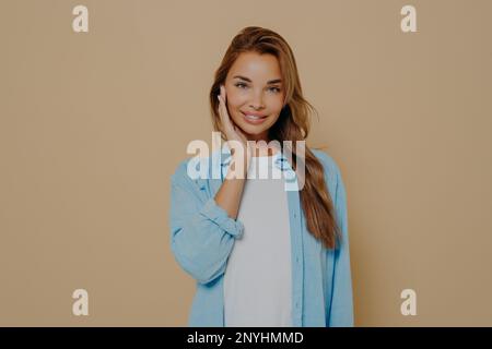 Portrait einer attraktiven, charmanten, brünetten Frau mit langen, rechten Haaren. Sie trägt ein blaues Hemd über einem weißen T-Shirt, lächelt in der Kamera, berührt ihre Wange Stockfoto