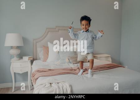 Fröhlicher kleiner gemischter Rennjunge glückliches Kind in blauem Hemd und beigefarbenen Shorts, das auf ein großes bequemes Bett mit Süßigkeiten springt und sein Glück mit Shinin ausdrückt Stockfoto