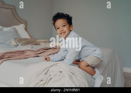 Süßer, glücklicher afroamerikanischer Junge, der spielt und Spaß auf dem Bett hat, nachdem er aus dem Kindergarten nach Hause gekommen ist, spielt und lächelt, während er Zeit im Bett verbringt Stockfoto