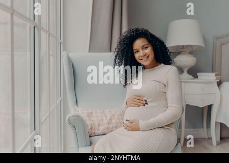 Glückliche junge, schwangere afro-amerikanische Dame in Kleid, die sich bald auf die Mutter vorbereitet, sitzt auf einem gemütlichen Sessel vor einem großen, geräumigen Fenster und smi Stockfoto
