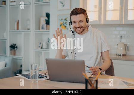 Lächelnder Freiberufler in Kopfhörern, winkte seinem Kollegen zu und hatte ein Online-Meeting, arbeitete von zu Hause aus, glücklicher Kerl, der auf die Laptop-Webcam schaute Stockfoto