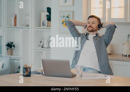 Fröhlicher, entspannter, bärtiger Mann, der Hände hinter dem Kopf hält und pausiert, während er online von zu Hause aus mit Headset und Laptop arbeitet, männlicher Freiberufler, der sich danach ausruht Stockfoto