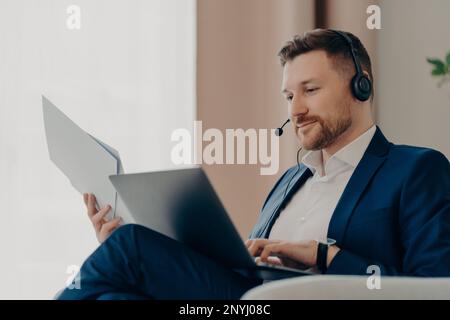 Geschäftsmann bei der Arbeit. Lächelnder, bärtiger männlicher Boss, der Dokumente und Laptop auf dem Schoß hielt, während er drinnen saß und ein Online-Meeting abhielt. Business-pe Stockfoto