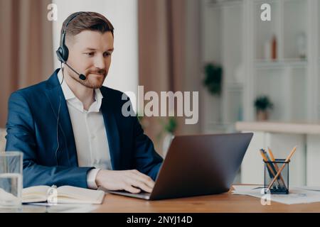 Aufmerksamer, junger, bärtiger Geschäftsmann im eleganten Anzug, der online telefoniert oder an einer Webkonferenz teilnimmt, während er ein Notebook und Kopfhörer mit Microp verwendet Stockfoto