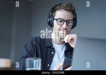 Junger fokussierter deutscher Mann mit Brille, der kabellose Kopfhörer mit Mobiltelefon trägt, am Arbeitsplatz im Büro Musik hört, lustige Internet-Videos ansieht Stockfoto