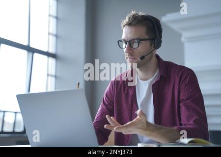 Europäischer Online-Lehrer mit Brille, sitzt am Schreibtisch in einem Headset und spricht per Videoanruf auf einem Laptop, während er von zu Hause aus arbeitet. Di Stockfoto