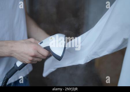 Zugeschnittenes Foto eines Mannes, der mit dem Dampfgarer ein weißes Hemd ordentlich bügelt, während er lange Ärmel hält. Geschäftsmann räumt formelle Kleidung vor wichtiger Gesellschaft auf Stockfoto