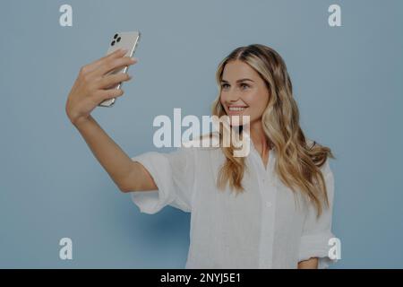 Blondes Teenager-Mädchen macht Selfie auf einem modernen Smartphone für Social-Media-Konten mit einem strahlenden Lächeln, lächelt für die Kamera und teilt Fotos mit ihrer Pommes Frites Stockfoto