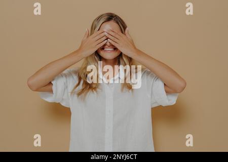 Glückliches, junges Mädchen mit blonden Haaren, das die Augen schließt, mit beiden Händen, mit aufgeregtem Gesichtsausdruck, lächelnd und wartend auf ein Geschenk zum Geburtstag, Cu Stockfoto