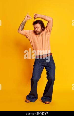 Studioaufnahme eines erwachsenen, freundlichen, bärtigen, haarigen Mannes, eines Hippies mit stilvoller Sonnenbrille und einer ausgestellten Jeans, die auf gelbem Hintergrund tanzen. Retro-Style Stockfoto