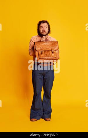 Porträt eines bärtigen, haarigen Mannes, schüchterner Wissenschaftler-Nerd in ausgestellten Jeans mit Aktentasche auf gelbem Hintergrund. Retro-Mode, amerikanisch Stockfoto