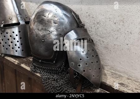 Der Helm aus Stahl eines mittelalterlichen Ritters mit Visier liegt auf einem rauen Holzregal vor der weißen Wand Stockfoto