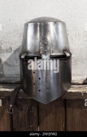 Ein geschlossener Helm aus Eisen mit einer Rüstung von Ritter und Ritter liegt auf einem rauen Holzregal vor der weißen Wand Stockfoto