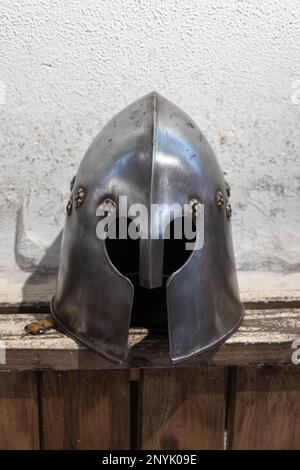 Der eiserne Helm des mittelalterlichen Ritters liegt auf einem rauen Holzregal vor der weißen Wand Stockfoto