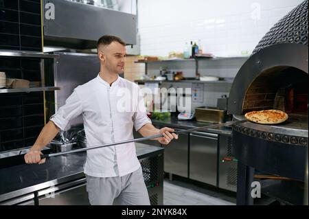 Junger männlicher Koch mit frisch gebackener Pizza auf langer Schaufel, selektiver Fokus Stockfoto