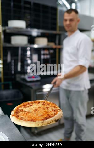 Junger männlicher Koch mit frisch gebackener Pizza auf langer Schaufel, selektiver Fokus Stockfoto
