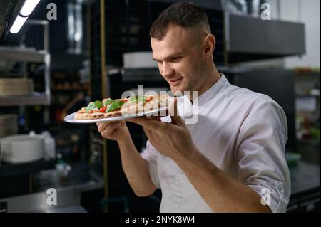 Zufriedener männlicher Koch genießt den Geruch von frisch gebackener Pizza Stockfoto