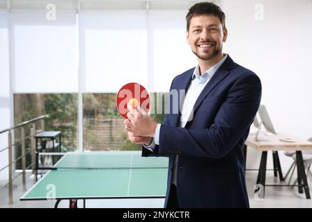 Geschäftsmann mit Tennisschläger und Ball in der Nähe des Tischtennistisches im Büro Stockfoto