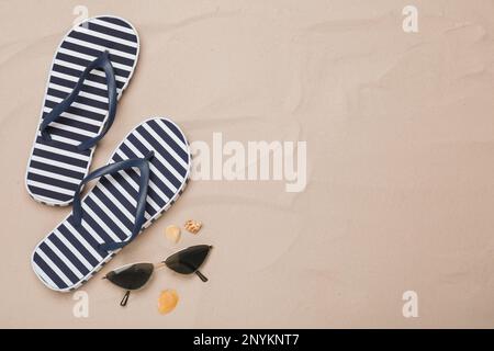 Gestreifte Flip-Flops, Sonnenbrille und Muscheln auf Sand, flach liegend. Platz für Text Stockfoto