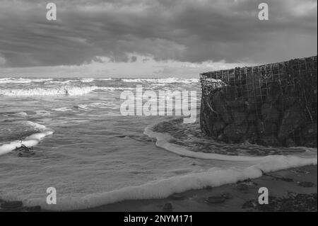 Gabionenkorb mit Steinen an einem Strand in Walton am Naze in Essex. Kalt, Winter, windiger Tag. Wellen schlagen gegen den Gabionenkorb. Stockfoto