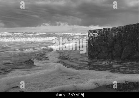 Gabionenkorb mit Steinen an einem Strand in Walton am Naze in Essex. Kalt, Winter, windiger Tag. Wellen schlagen gegen den Gabionenkorb. Stockfoto