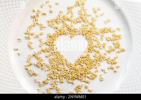 Chaotische Nudeln kleine Buchstaben und Ziffern in der Form eines Herzens auf einem weißen Teller verstreut. Ungekocht Trockenfutter Zutat Hintergrund. Liebe für cooki Stockfoto