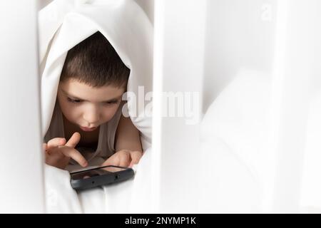 Ein 5-jähriger Junge spielt Spiele auf seinem Handy, während er im Bett liegt. Geräte im Bett vor dem Schlafengehen bei einem Kind Stockfoto