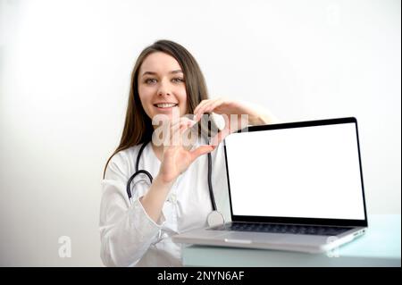 Arzthelferin in der Nähe des Computers mit weißem Bildschirm Leerraum für Text zeigt mit Händen lächelndes Herz Nahrungsergänzungsmittel gute Medizin gegenseitige Hilfe ausgezeichnete Behandlung Medizin Produktwerbung Stockfoto