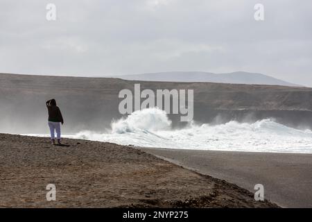 Eine Dame beobachtet starke Wellen, die aus der Ferne auf Felsen krachen. Ajuy, Fuerteventura. Stockfoto