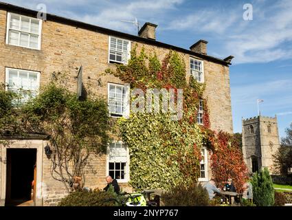 Die Gäste sitzen an einem sonnigen Herbsttag vor dem Rose and Crown Hotel in Romaldkirk Village. Mauern mit Efeu und virginia Creeper. Hinter der Kirche St. Romalds. Stockfoto