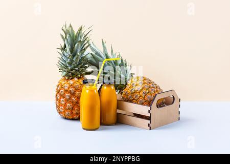 Ananas in einer Holzschachtel und ein frischer, gesunder Smoothie in Glasflaschen und einem Strohhalm auf blauem Hintergrund. Gesund, Entgiftung und diätetische Ernährung conce Stockfoto