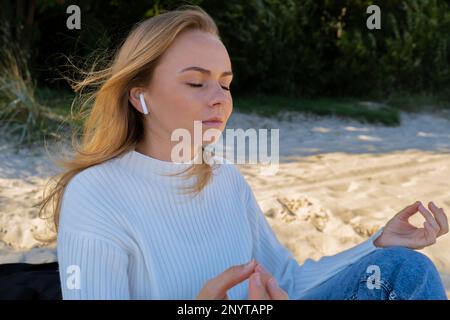 Glückliche junge Frau am Strand Meer Meer. Tragen von kabellosen Kopfhörern, die eine Audio-Heilklang-Therapie durchführen, während Sie sich der Natur bewusst sind. Wohlbefinden Einheit mit Natur Gesundheit Achtsamkeit. Genießen Sie Entspannung im Freien Stockfoto