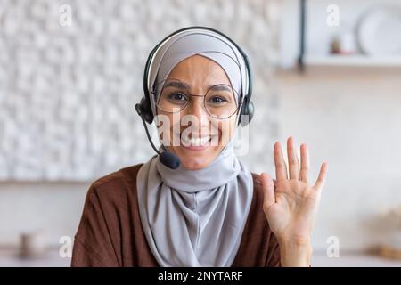 Nahaufnahme. Porträt einer jungen Studentin in einem Hidschab, einer Brille und einem Headset. Er sitzt und lächelt vor der Kamera, winkt mit der Hand, sagt Hallo. Stockfoto