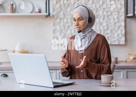 Online-Schulung. Eine junge muslimische Lehrerin in einem Hidschab, einer Brille und einem Headset. Er sitzt zu Hause in der Küche vor seinem Laptop und plappert, unterrichtet, unterrichtet per Videoanruf. Stockfoto